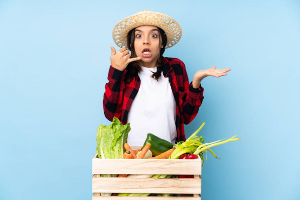 Joven agricultora Mujer sosteniendo verduras frescas en una canasta de madera haciendo gesto telefónico y dudando - Foto, imagen