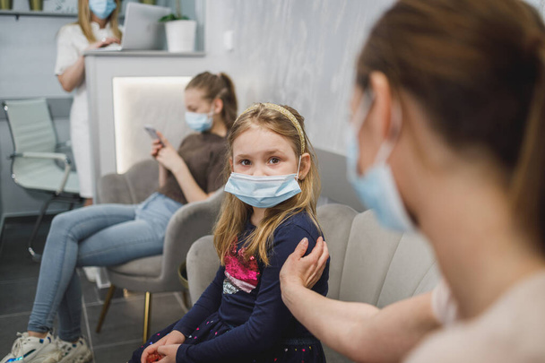 Pensive little girl and her mom with face mask in waiting room at dentist's office. - Foto, Imagem