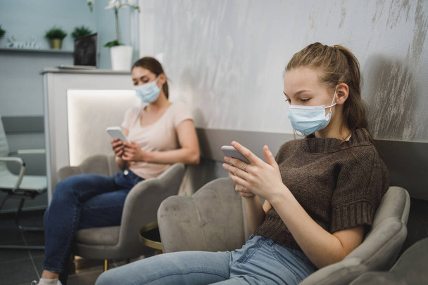 Una ragazza carina adolescente indossando maschera facciale e usando il suo cellulare in sala d'attesa presso lo studio del dentista. - Foto, immagini