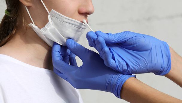 рука врача, выполняющего анализ носовой полости на обнаружение коронавируса молодой девушке в белой хирургической маске - Фото, изображение