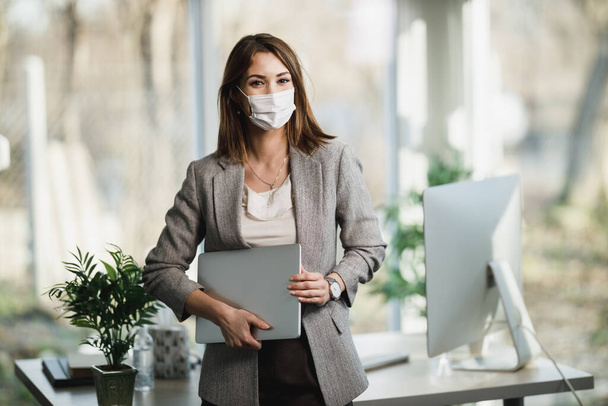 Młoda, odnosząca sukcesy biznesmenka z maską chirurgiczną trzymająca laptopa i stojąca w biurze. Patrzę w kamerę.. - Zdjęcie, obraz