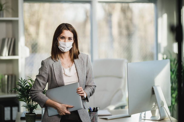 Een succesvolle jonge zakenvrouw met een chirurgisch masker die laptop vasthoudt en in haar kantoor staat. Kijkend naar camera. - Foto, afbeelding