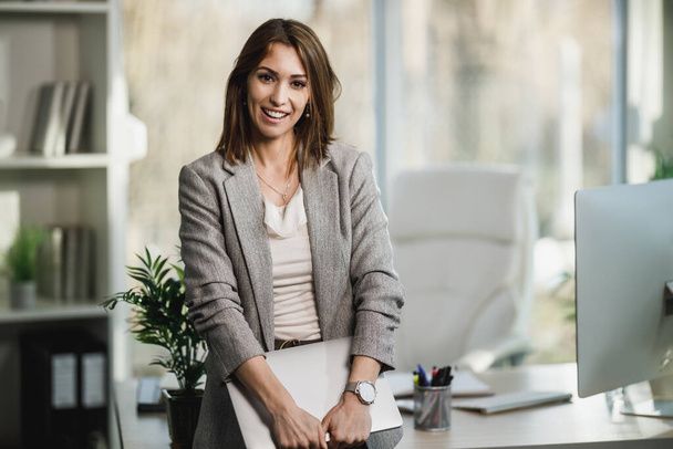 Μια επιτυχημένη νεαρή επιχειρηματίας που κρατάει λάπτοπ και στέκεται στο γραφείο της. Κοιτάζοντας την κάμερα. - Φωτογραφία, εικόνα