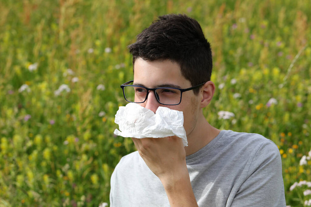 mladý krátkozraký chlapec s brýlemi kýchnout, protože alergie na trávu na květinovém poli na jaře - Fotografie, Obrázek