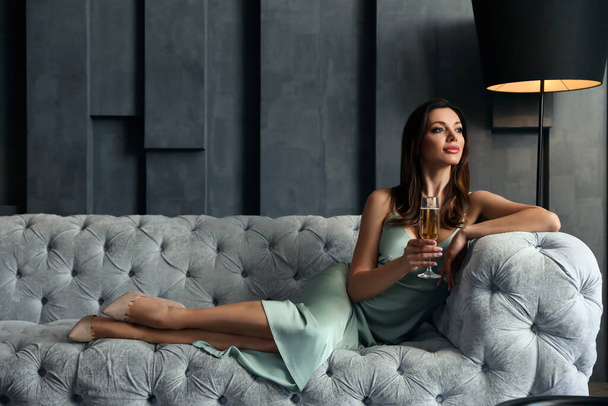 Beautiful woman in elegant dress with glass of champagne on sofa indoors. Luxury lifestyle - Foto, Bild