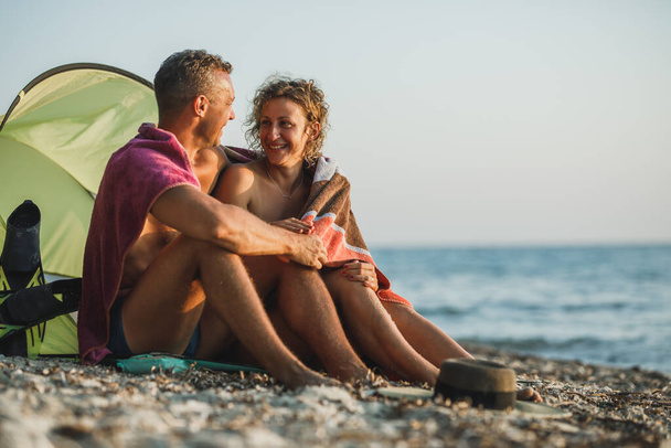 Una joven pareja cariñosa pasando el día en la playa y disfrutando de la puesta de sol envuelta en una manta. - Foto, Imagen