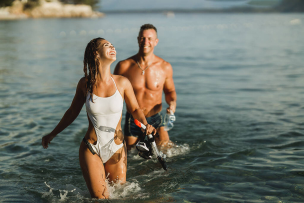Una pareja alegre con sus máscaras de buceo y snorkels caminando por el mar después de bucear. - Foto, Imagen