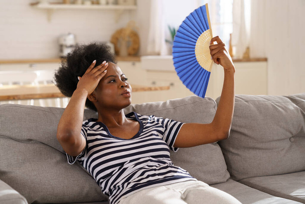 Μαύρη γυναίκα που υποφέρει από θερμοπληξία επίπεδη χωρίς air-condition κυματίζει ανεμιστήρα που βρίσκεται στον καναπέ στο σπίτι - Φωτογραφία, εικόνα