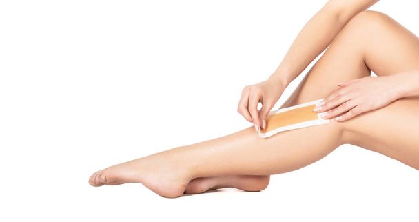Beautician waxing female legs in spa center. Well-groomed woman legs after depilation isolated on white background. Waxing woman leg with wax strip at beauty spa. Close-up waxing woman leg in spa - 写真・画像
