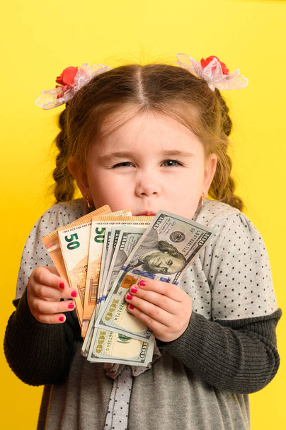 Niña caucásica sosteniendo dinero, retrato de un niño sobre un fondo amarillo, educación financiera de los niños. nuevo - Foto, Imagen
