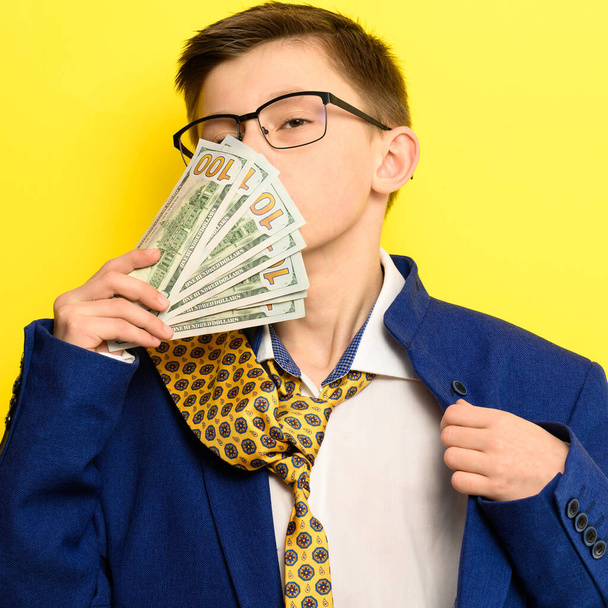 Un tipo con un gran traje clásico sobre un fondo amarillo sostiene una moneda extranjera, un niño con gafas y un traje con corbata. nuevo - Foto, imagen
