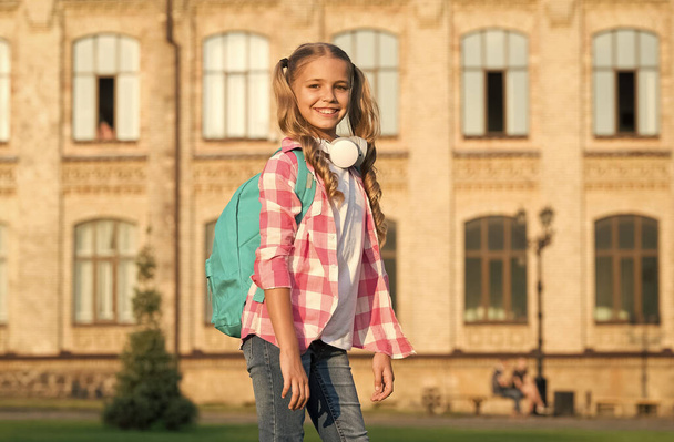 small girl wear checkered shirt. child with headset at sunset. kid spring and summer fashion. happy childhood. e-learning with ebook. modern technology. cheerful music lover with backpack - Valokuva, kuva