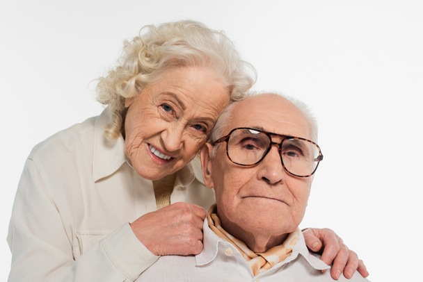 ευτυχισμένο ηλικιωμένο ζευγάρι αγκαλιάζει απαλά και κοιτάζοντας κάμερα απομονωμένη σε λευκό - Φωτογραφία, εικόνα