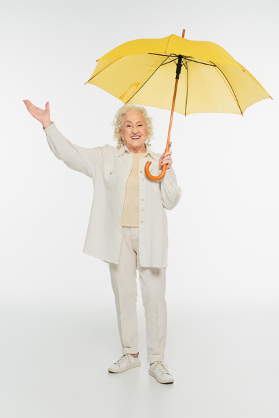 smiling elderly woman in casual clothes with triumph gesture standing with yellow umbrella on white - 写真・画像