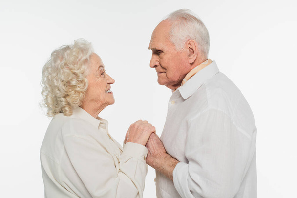 happy elderly couple gently holding hands and looking at each other isolated on white - Photo, Image