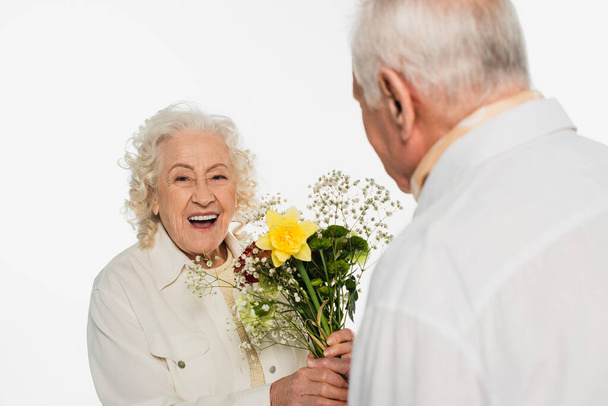 smiling elderly woman holding bouquet of flowers near blurred husband isolated on white - Foto, Imagem
