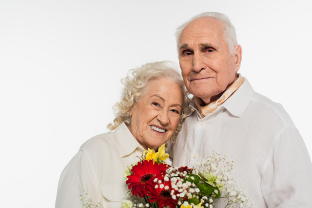 幸せそうなお年寄りカップルが花束を持って白で孤立したカメラを見て - 写真・画像