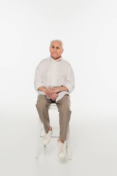 λυπημένος ηλικιωμένος άνδρας με καθημερινά ρούχα κάθεται σε σκαμνί σε λευκό - Φωτογραφία, εικόνα
