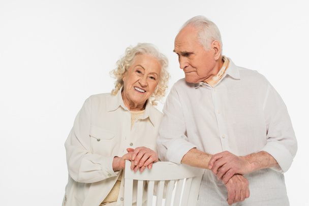 felice coppia anziana in abiti casual in piedi vicino sgabello isolato su bianco - Foto, immagini