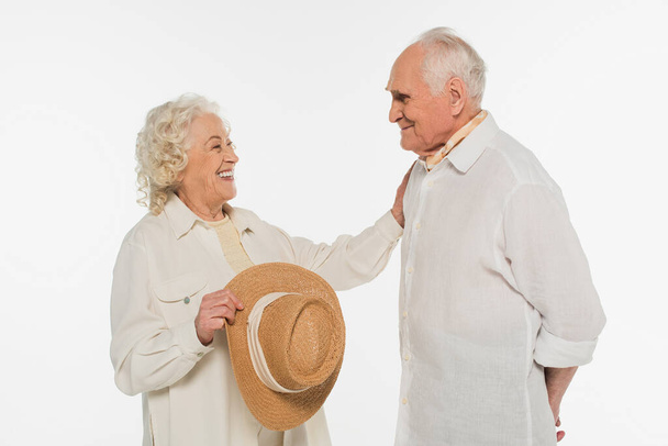 sonriente anciana sosteniendo sombrero y tocando marido con la mano extendida aislada en blanco - Foto, Imagen