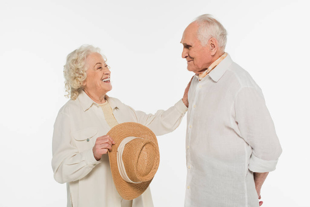 positive elderly woman holding hat and touching husband with outstretched hand isolated on white - Fotoğraf, Görsel