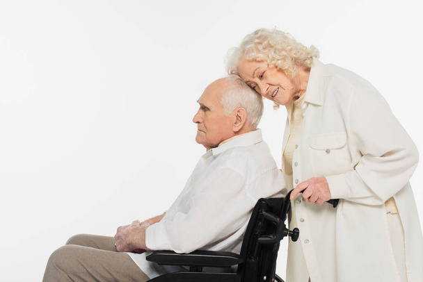 oudere vrouw zachtjes aanraken echtgenoot in rolstoel met hoofd geïsoleerd op wit - Foto, afbeelding