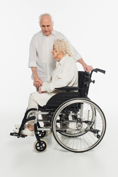 smutna starsza kobieta na wózku inwalidzkim trzymając męża za rękę na białym - Zdjęcie, obraz