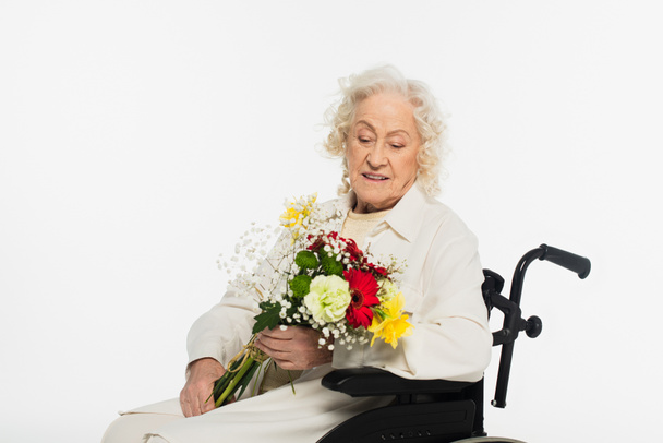 anciana sonriente en silla de ruedas sosteniendo ramo de flores aisladas en blanco - Foto, imagen