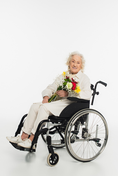 smiling elderly woman in casual clothes sitting in wheelchair holding bouquet of flowers on white - Photo, Image