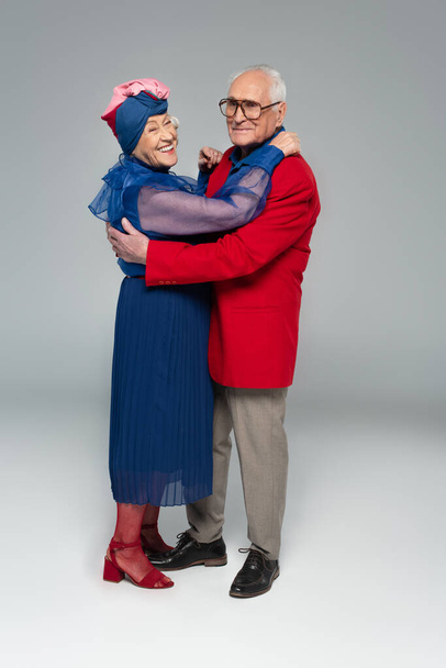 sonriente pareja de ancianos en vestido azul y chaqueta roja abrazándose en gris - Foto, imagen