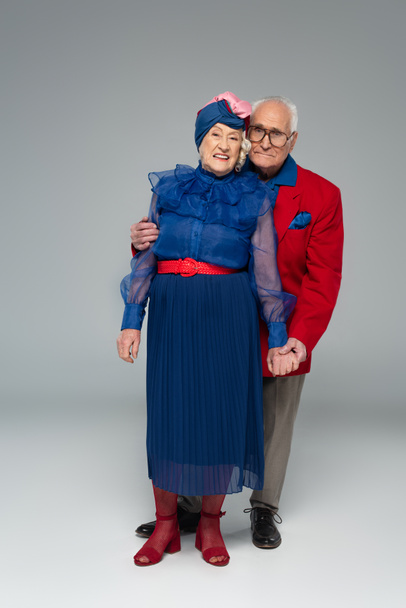 couple âgé souriant en robe bleue et blazer rouge étreignant et tenant la main sur gris - Photo, image