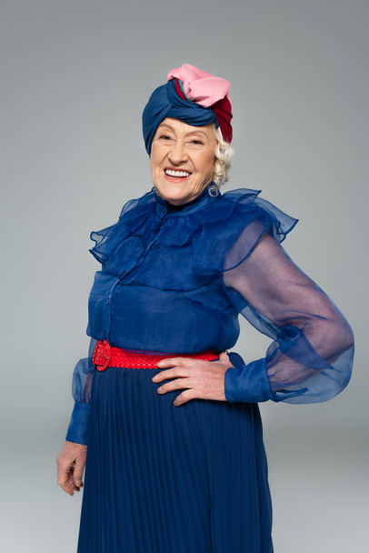 smiling elderly woman in blue dress and turban posing with hand on hip isolated on grey - Foto, imagen