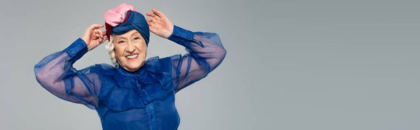 smiling elderly woman in blue dress adjusting turban with hands isolated on grey, banner - Фото, изображение