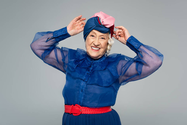 smiling elderly woman in blue dress adjusting turban with hands isolated on grey - Φωτογραφία, εικόνα