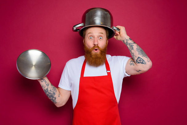 se preguntaba chef con barba y delantal rojo juega con olla - Foto, imagen