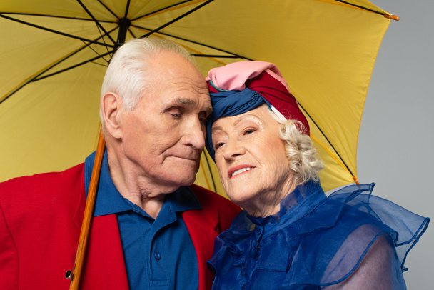 elderly man with yellow umbrella hugging wife in blue dress and turban isolated on grey - Fotó, kép