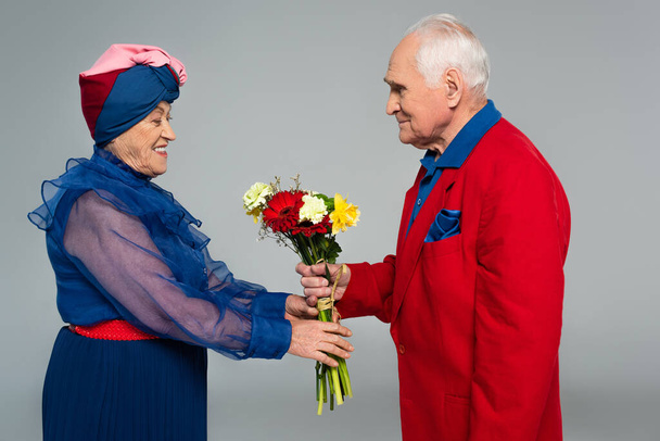 usmívající se starší muž v červeném blejzru představující kytice květin na manželku v modrých šatech a turban izolované na šedé - Fotografie, Obrázek