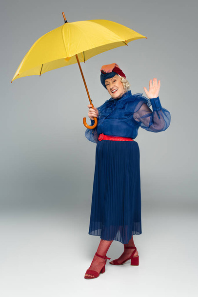 Lächelnde ältere Frau in blauem Kleid und Turban mit gelbem Regenschirm und winkend mit der Hand auf grau - Foto, Bild