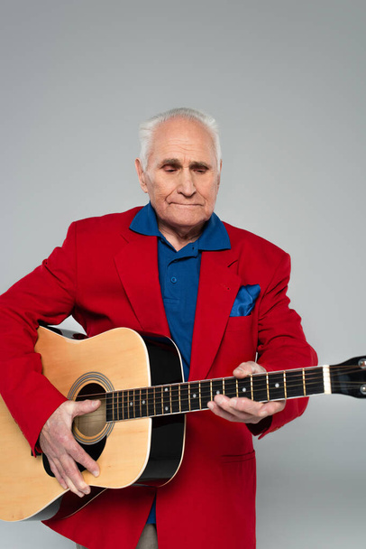 elderly man in red blazer holding acoustic guitar isolated on grey - Foto, afbeelding