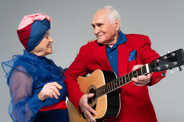Lächelnder älterer Mann im roten Blazer spielt Akustikgitarre neben tanzender Frau in blauem Kleid und Turban isoliert auf grau - Foto, Bild
