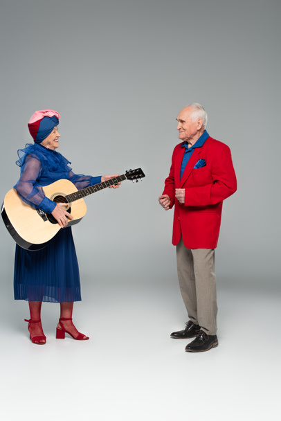 positiivinen iäkäs pari tyylikkäissä vaatteissa tanssii akustisen kitaran kanssa harmaalla - Valokuva, kuva