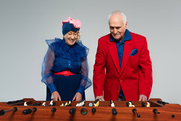 positive elderly couple in stylish clothes playing table football on grey - Фото, изображение