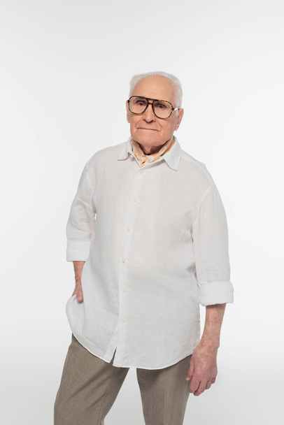 smiling elderly man in casual clothes and glasses posing with hand in pocket and looking at camera isolated on white - Foto, immagini
