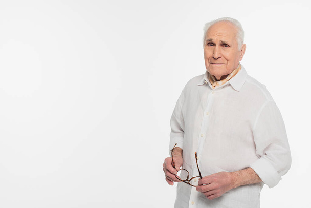sonriente anciano sosteniendo gafas y mirando a la cámara aislada en blanco - Foto, Imagen