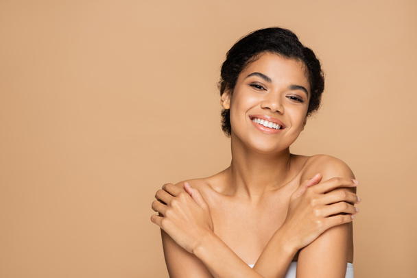 happy african american woman touching bare shoulders and looking at camera isolated on beige  - Photo, Image