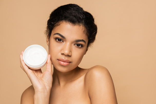 african american woman with bare shoulders holding container with face cream isolated on beige  - Photo, Image