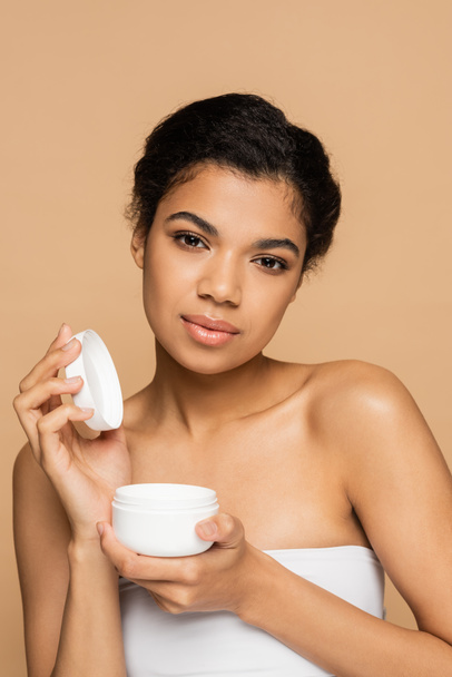 african american woman with bare shoulders holding container with face cream isolated on beige  - Фото, изображение