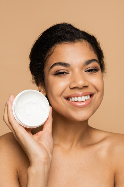 pleased african american woman with bare shoulders holding container with face cream isolated on beige  - Фото, зображення