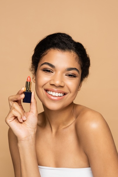 happy african american woman with bare shoulders holding lipstick tube isolated on beige  - Foto, imagen