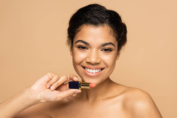 alegre mujer afroamericana con hombros desnudos sosteniendo lápiz labial aislado en beige  - Foto, imagen